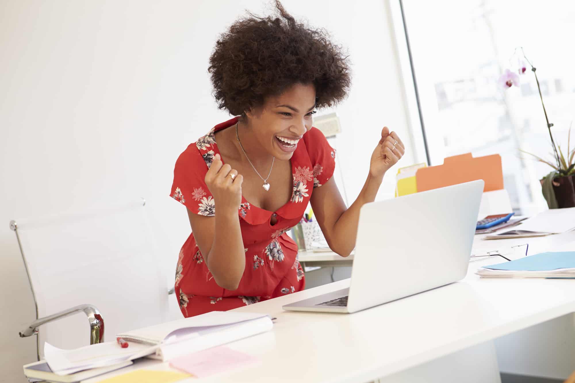 Photo shows a happy, excited woman standing behind her desk, looking at her laptop screen. This is the featured image for a blog post about free blogging tools to help make your posts shine.