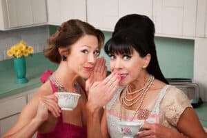 Photo of two women dressed in retro 1950s outfits. They're in a kitchen, sipping tea and one woman is whispering a secret to the other. The purpose of the photo is to make you think of etiquette because the post is about the rules of social media and why etiquette matters.