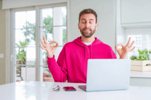 Young man with beard meditating at his desk. He is wearing a hot pink hoodie and the desk and surrounding area are very clean and tidy. There is a laptop, phone and pen on the desk, neatly arranged. This is the featured image for a blog post titled "How to Marie Kondo Your Nonprofit Website So Great Content Can Shine