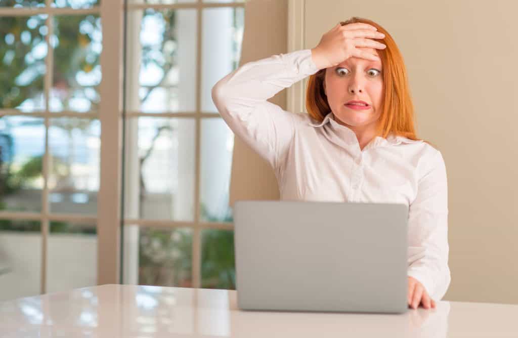 Mid 30s caucasian woman with long read hair, wearing a white shirt. She is stressed out, with one hand slapped against her forehead as she looks down at her laptop screen. This is the featured image for an article about Website Design Mistakes.